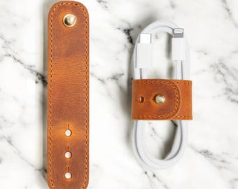 May include: Two brown leather cable organizers with brass hardware. One is a long, rectangular strap with three holes. The other is a small loop securing a white charging cable. The leather has a distressed finish.