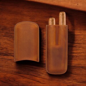 May include: Two brown leather cigar cases, one with two cigars inside and the other empty. The cases are on a wooden surface.