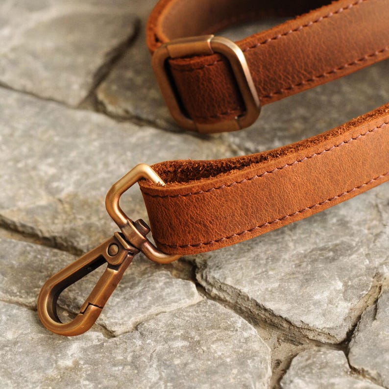 May include: A brown leather strap with a brass buckle and a brass hook. The strap is stitched with a brown thread.