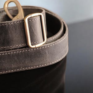 May include: Close-up of a gray leather strap with a brass buckle. The strap is stitched with a light gray thread.
