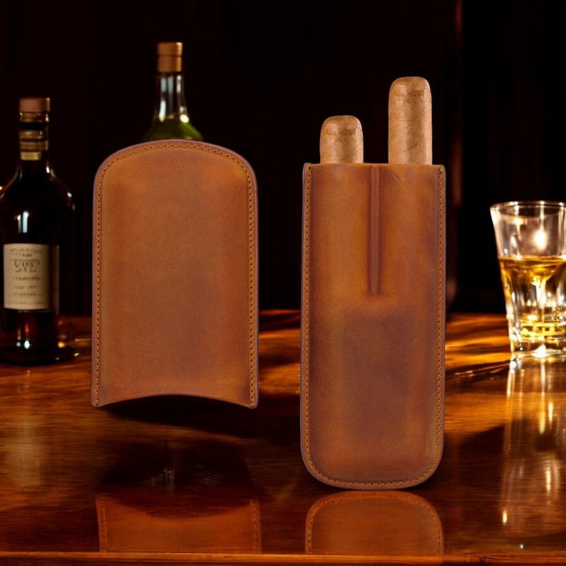 May include: Two brown leather cigar cases with a stitched edge. The case on the right has two cigars inside.