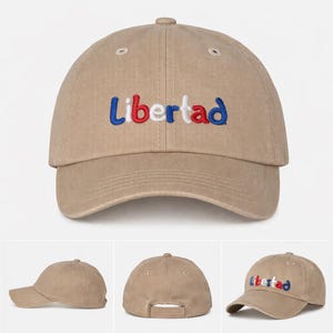 May include: A tan baseball cap with the word "Libertad" embroidered in blue, red, and white. The cap has a curved brim and an adjustable strap at the back. The hat is made of a soft, durable fabric.