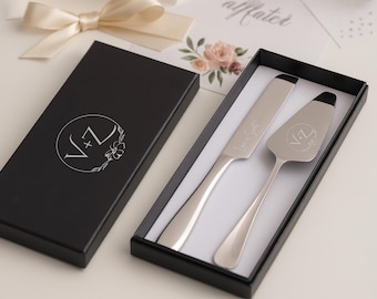 May include: A silver cake knife and server set in a black box. The knife and server have black accents and are engraved with initials. The box has a white floral design and initials on the lid. A cream-colored bow is in the background.