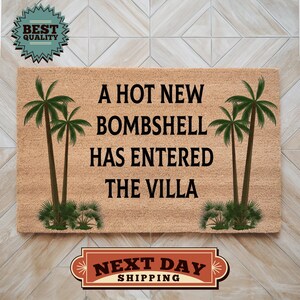 May include: A rectangular tan coir doormat featuring the black text A HOT NEW BOMBSHELL HAS ENTERED THE VILLA. The design includes two green palm trees on the left side and two green palm trees on the right side of the central text.