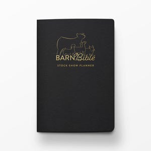 May include: A black notebook with the words "BARN Bible STOCK SHOW PLANNER" in gold lettering. The cover features a gold outline of a cow and three smaller animals. The notebook is closed and has a textured surface.