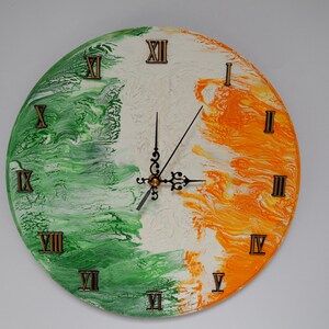 Peut inclure: Une horloge murale ronde avec un motif de peinture marbrée en vert, blanc et orange. Le cadran affiche des chiffres romains en relief de couleur bronze pour chaque heure et comprend deux aiguilles en métal noir ornées au centre.