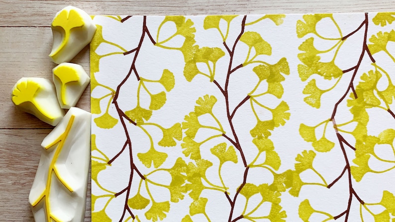 A hand carved rubber stamp set of ginkgo tree design - 3 ginkgo leaves and 1 tree branch, shown with a stamped example in yellow green and brown on white paper. Close-up view of stamped example.