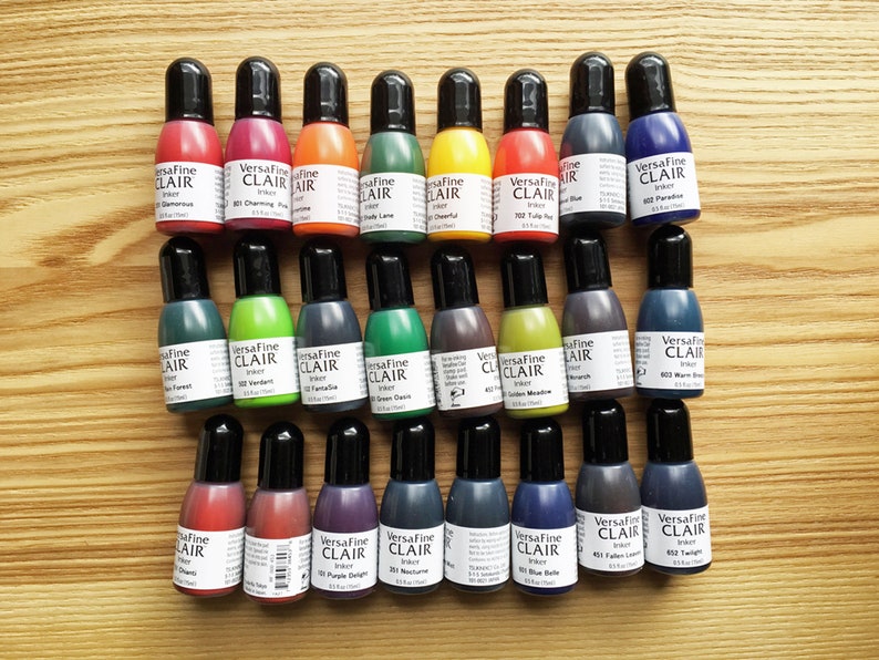 K&ouml;nnte beinhalten: Eine Sammlung von 18 kleinen Flaschen VersaFine Clair Tinte in verschiedenen Farben, darunter Rot, Orange, Gelb, Gr&uuml;n, Blau, Lila und Braun. Die Flaschen sind in drei Reihen mit jeweils sechs Flaschen angeordnet. Jede Flasche hat einen schwarzen Verschluss und ein Etikett mit dem VersaFine Clair Logo und dem Namen der Tintenfarbe.