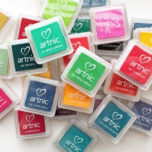 Puede incluir: Una colecci&oacute;n de peque&ntilde;os contenedores cuadrados de pl&aacute;stico transparente con almohadillas de tinta de colores en su interior. Cada almohadilla tiene un logotipo en forma de coraz&oacute;n y la palabra "artnic" impresa en ella. Las almohadillas de tinta est&aacute;n etiquetadas con n&uacute;meros y nombres de colores.