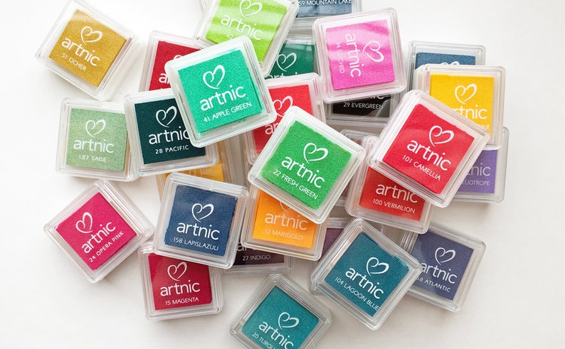 Puede incluir: Una colecci&oacute;n de peque&ntilde;os contenedores cuadrados de pl&aacute;stico transparente con almohadillas de tinta de colores en su interior. Cada almohadilla tiene un logotipo en forma de coraz&oacute;n y la palabra "artnic" impresa en ella. Las almohadillas de tinta est&aacute;n etiquetadas con n&uacute;meros y nombres de colores.