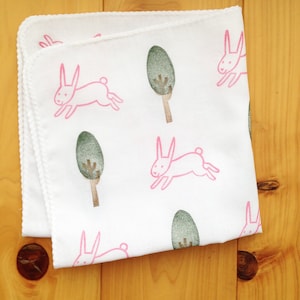 May include: White cotton baby blanket with a pink bunny rabbit and green tree design.