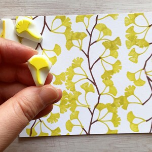 A hand carved rubber stamp set of ginkgo tree design - 3 ginkgo leaves and 1 tree branch, shown with a stamped example in yellow green and brown on white paper. One of the stamps is held in the hand to give an idea how big it is.
