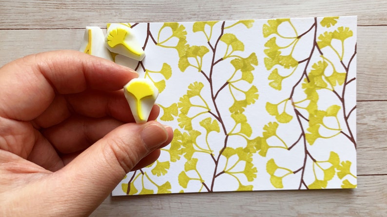 A hand carved rubber stamp set of ginkgo tree design - 3 ginkgo leaves and 1 tree branch, shown with a stamped example in yellow green and brown on white paper. One of the stamps is held in the hand to give an idea how big it is.