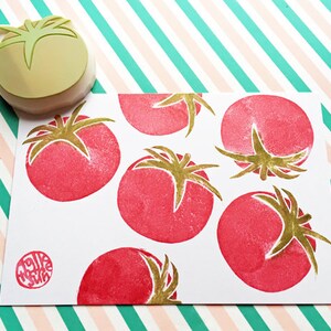Pode incluir: Um cart&atilde;o branco com um padr&atilde;o de tomate vermelho e dourado. O cart&atilde;o tem um pequeno carimbo redondo vermelho com o texto "Make Wish" em branco.
