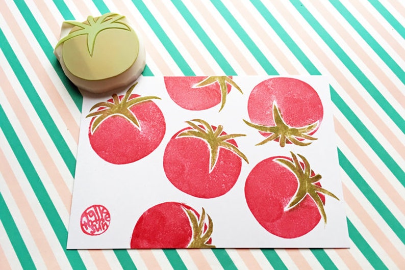 Pode incluir: Um cart&atilde;o branco com um padr&atilde;o de tomate vermelho e dourado. O cart&atilde;o tem um pequeno carimbo redondo vermelho com o texto "Make Wish" em branco.