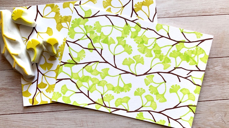 A hand carved rubber stamp set of ginkgo tree design - 3 ginkgo leaves and 1 tree branch, shown with a stamped example in yellow green and brown on white paper.