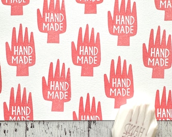 May include: A white background with a repeating pattern of red handprints that say "HAND MADE" in white text. A white rubber stamp with the same design is shown in the lower right corner.