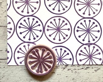 May include: A close-up of a purple rubber stamp with a circular design of a starburst with a white background. The stamp is on top of a sheet of paper with a repeating pattern of the same starburst design in purple.