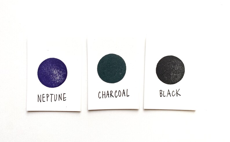 Puede incluir: Tres muestras de pintura en tarjetas blancas. Las muestras est&aacute;n etiquetadas como "Neptuno", "Carb&oacute;n vegetal" y "Negro". Las muestras son de color morado oscuro, verde oscuro y negro.