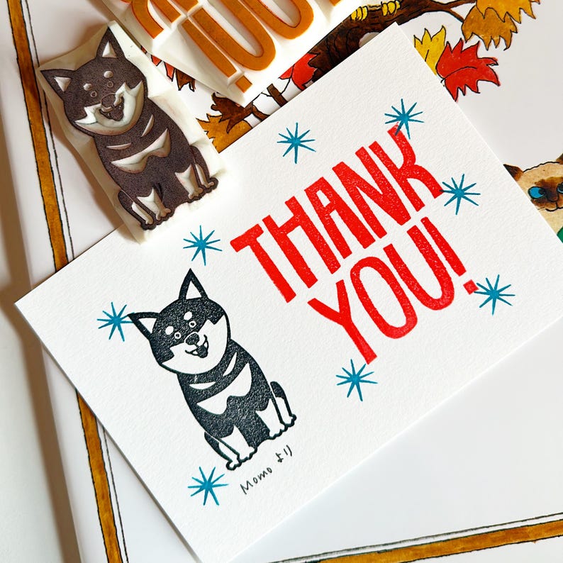 A sample image of handmade postcard using hand carved rubber stamps available in the shop. The text THANK YOU in red and a black Shiba Inu in black with stars in blue printed around the card.