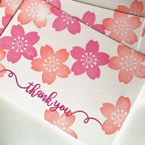 A sample image of thank you cards created with hand carved rubber stamps available in the shop. Cherry blossoms and a message "thank you" are stamped in the white card in coral pink and cherry pink.