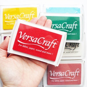 Pode incluir: Um almofada de tinta vermelha com o texto "VersaCraft" e as palavras "Para tecido, papel, madeira e mais!" O almofada de tinta est&aacute; em uma caixa de pl&aacute;stico transparente e est&aacute; sendo segurado em uma m&atilde;o.