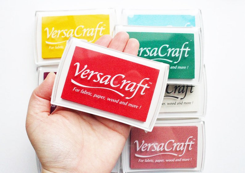 Pode incluir: Um almofada de tinta vermelha com o texto "VersaCraft" e as palavras "Para tecido, papel, madeira e mais!" O almofada de tinta est&aacute; em uma caixa de pl&aacute;stico transparente e est&aacute; sendo segurado em uma m&atilde;o.