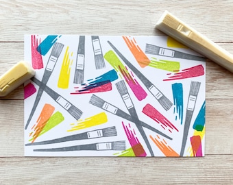 Puede incluir: Una tarjeta blanca con un patrón repetitivo de pinceles coloridos. Los pinceles son de varios colores, incluyendo rojo, naranja, amarillo, verde, azul y morado. La tarjeta está rodeada por una superficie de madera.
