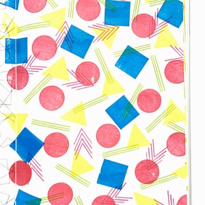 May include: A white background is covered with a repeating pattern of pink circles, blue squares, yellow triangles, and green and pink lines. The shapes are arranged in a playful, geometric design.