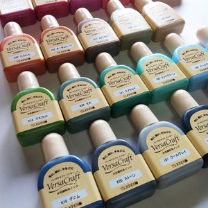 May include: A collection of 18 small bottles of VersaCraft water-based ink in various colors. The bottles are labeled with the color name and the brand name.