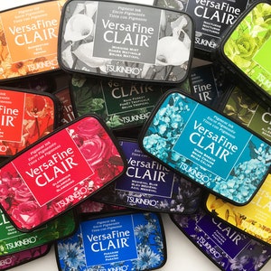 May include: A collection of VersaFine CLAIR pigment ink pads in various colors, each with floral designs and the brand name. The ink pads are rectangular and feature the text "VersaFine CLAIR" and the ink color name.
