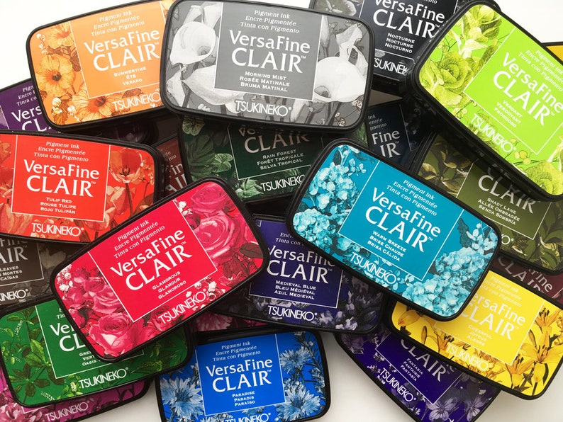 May include: A collection of VersaFine CLAIR pigment ink pads in various colors, each with floral designs and the brand name. The ink pads are rectangular and feature the text "VersaFine CLAIR" and the ink color name.