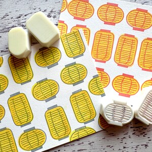 May include: Four white rubber stamps with a raised design of a Japanese lantern. The stamps are on a white surface with two pieces of paper with a repeating pattern of yellow and orange lanterns.