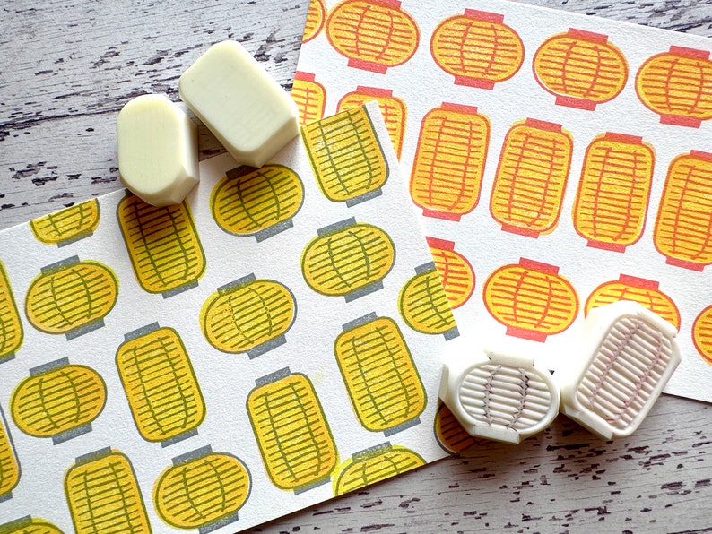 May include: Four white rubber stamps with a raised design of a Japanese lantern. The stamps are on a white surface with two pieces of paper with a repeating pattern of yellow and orange lanterns.