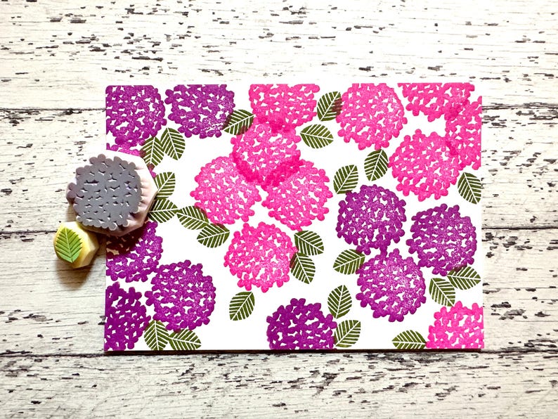 A hand carved rubber stamp set of hydrangea design - flower and leaf, shown with a stamped example in various colored random pattern on white paper.