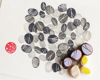 May include: A collection of gray and white patterned rubber stamps in the shape of pebbles. The stamps are arranged in a loose circle on a white background. There are also five smaller rubber stamps in the shape of pebbles in purple, brown, and yellow.