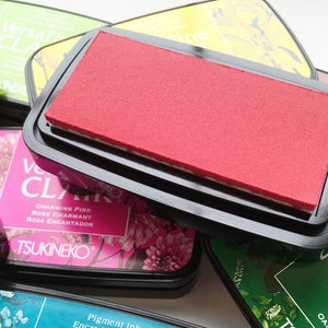 May include: A collection of VersaFine Clair pigment ink pads in various colors, including red, green, and pink. The pads are in black plastic cases, with the red pad prominently displayed. Floral designs and text are visible on the labels.