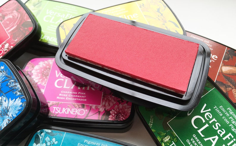May include: A collection of VersaFine Clair pigment ink pads in various colors, including red, green, and pink. The pads are in black plastic cases, with the red pad prominently displayed. Floral designs and text are visible on the labels.