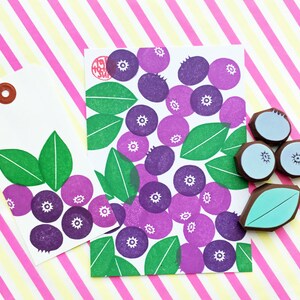 K&ouml;nnte beinhalten: Zwei wei&szlig;e Geschenkanh&auml;nger mit einem Muster aus lila und gr&uuml;nen Blaubeeren. Ein Anh&auml;nger hat einen braunen Kreis f&uuml;r eine Aufh&auml;ngeschlaufe. Der andere Anh&auml;nger liegt auf einem rosa und gelben gestreiften Hintergrund. Es gibt auch drei blaue Stempel mit Blaubeeren und einen gr&uuml;nen Blattstempel.