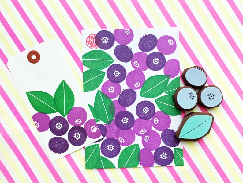 K&ouml;nnte beinhalten: Zwei wei&szlig;e Geschenkanh&auml;nger mit einem Muster aus lila und gr&uuml;nen Blaubeeren. Ein Anh&auml;nger hat einen braunen Kreis f&uuml;r eine Aufh&auml;ngeschlaufe. Der andere Anh&auml;nger liegt auf einem rosa und gelben gestreiften Hintergrund. Es gibt auch drei blaue Stempel mit Blaubeeren und einen gr&uuml;nen Blattstempel.