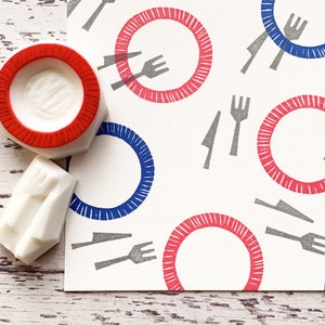 May include: Two white rubber stamps, one shaped like a plate with a red rim and the other shaped like a fork and knife, are next to a white card with a repeating pattern of red and blue plates with gray forks and knives.