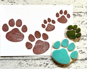 Puede incluir: Cuatro sellos de goma con huellas de patas. Los sellos son de diferentes tamaños y colores, incluyendo marrón, verde y azul.