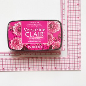 May include: A rectangular ink pad with a pink floral design and the text "VersaFine CLAIR" in white. The ink pad is labeled "Charming Pink" and measures approximately 3 inches by 2 inches.