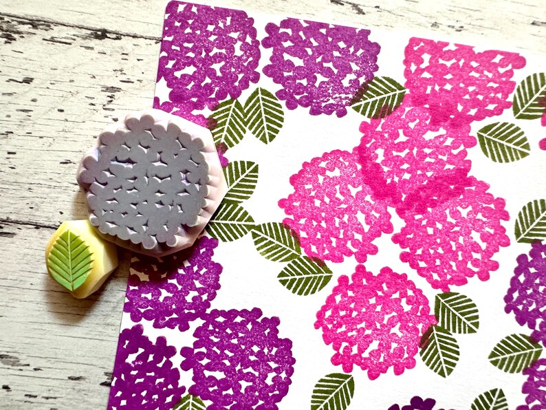 A hand carved rubber stamp set of hydrangea design - flower and leaf, shown with a stamped example in various colored random pattern on white paper.