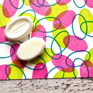 May include: Two white rubber stamps with oval shapes, one with a textured surface, on a white background with a pattern of overlapping pink, green, and blue ovals.