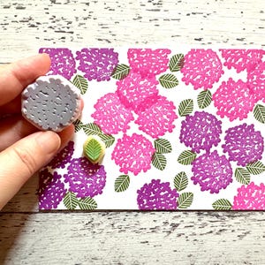 A hand carved rubber stamp set of hydrangea design - flower and leaf, shown with a stamped example in various colored random pattern on white paper.
One of the stamps is held in the hand to give an idea of its size.