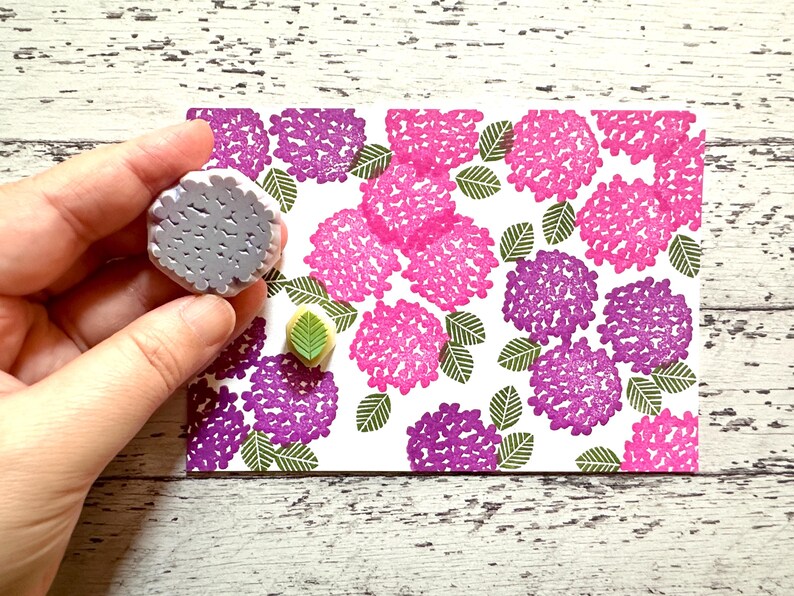 A hand carved rubber stamp set of hydrangea design - flower and leaf, shown with a stamped example in various colored random pattern on white paper.
One of the stamps is held in the hand to give an idea of its size.