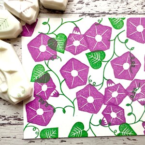 May include: A white card with a repeating pattern of purple morning glory flowers and green leaves. The flowers are stylised and have a white centre. The leaves are simple and have a textured look.
