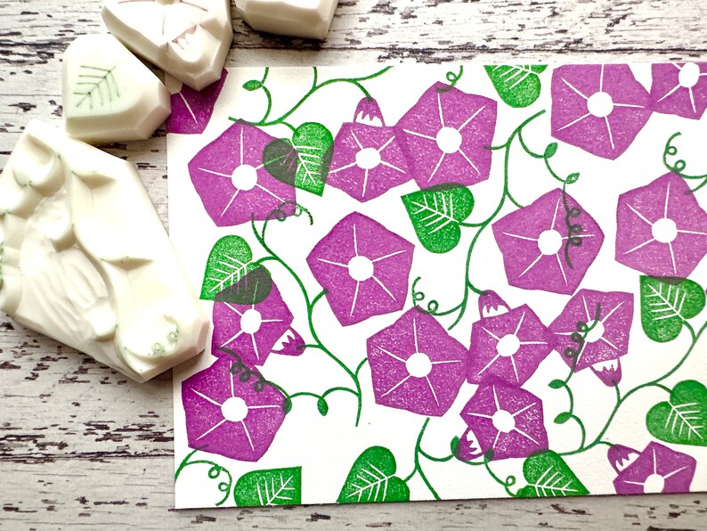 May include: A white card with a repeating pattern of purple morning glory flowers and green leaves. The flowers are stylised and have a white centre. The leaves are simple and have a textured look.