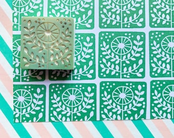 May include: A green rubber stamp with a white design of a stylized flower with leaves. The stamp is on a green and white patterned background.